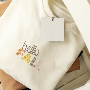 Puede incluir: Una sudadera blanca con el texto "hello fall" bordado en rayas de colores. La sudadera est&aacute; doblada y se muestra en un estante blanco.