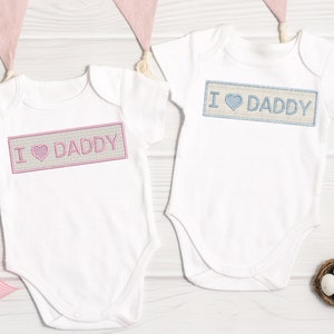 May include: Two white baby onesies with short sleeves. The onesies have the words "I love Daddy" embroidered on them. One onesie has pink embroidery and the other has blue embroidery.