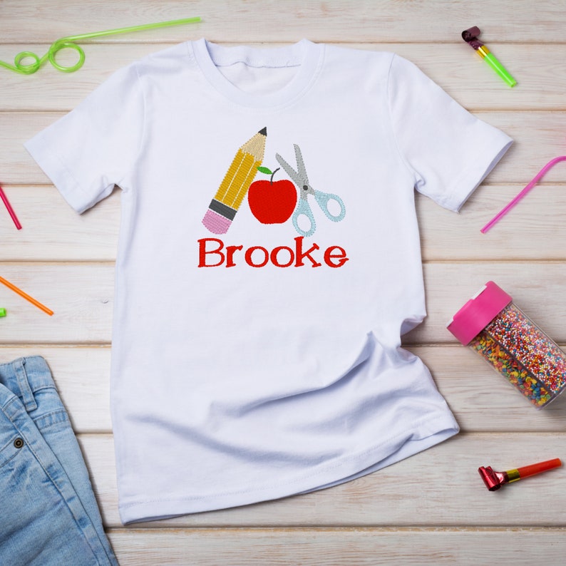May include: White t-shirt with a red apple, yellow pencil, and silver scissors embroidered on the front. The name "Brooke" is embroidered below the school supplies.