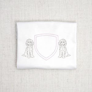 May include: White fabric with a shield design and two dog illustrations. The shield is outlined in a light purple color. The dogs are drawn in a detailed, sketch-like style. The fabric is folded and appears to be a towel or cloth.