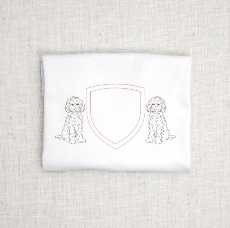 May include: White fabric with a shield design and two dog illustrations. The shield is outlined in a light purple color. The dogs are drawn in a detailed, sketch-like style. The fabric is folded and appears to be a towel or cloth.
