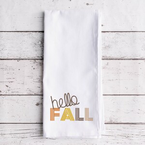 Puede incluir: Toalla de cocina blanca con el texto "hello fall" bordado en marr&oacute;n, amarillo y naranja.