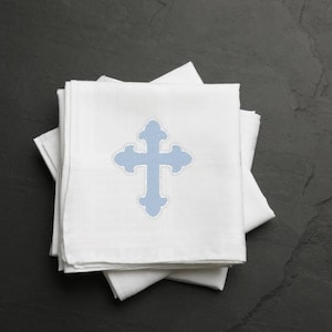 Könnte beinhalten: Eine weiße Stoffserviette mit einem blauen Kreuz, das darauf gestickt ist. Das Kreuz hat ein wellenförmiges Muster.