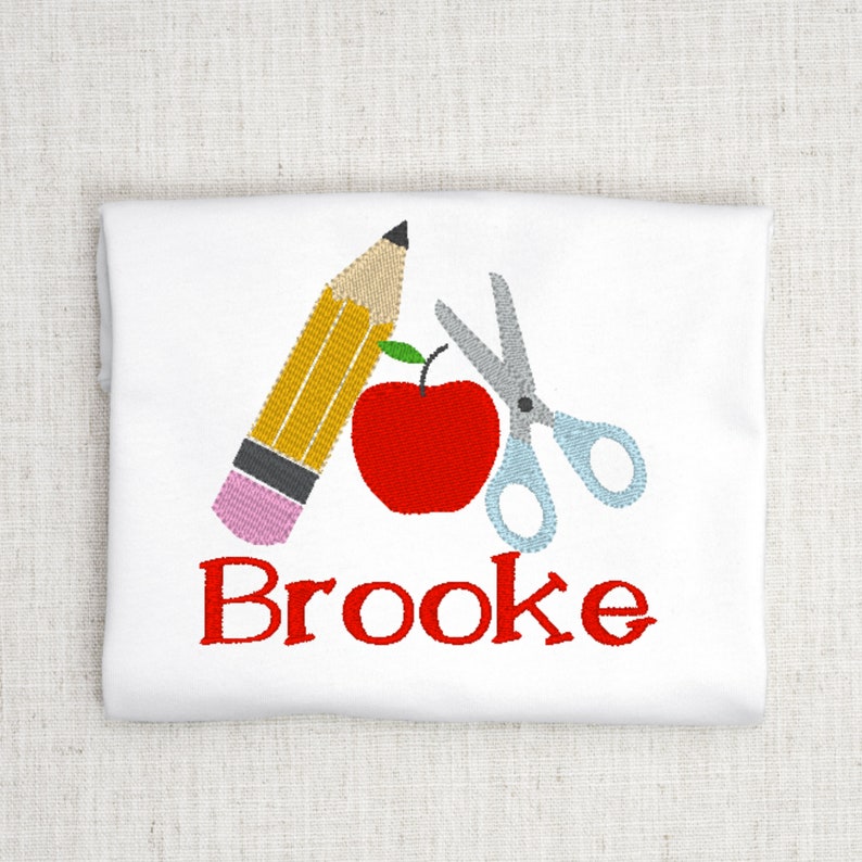May include: White t-shirt with an embroidered design of a red apple, a yellow pencil, and a pair of silver scissors. The name "Brooke" is embroidered in red below the design.