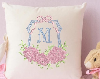 May include: A white pillow with a blue and pink embroidered monogram design. The monogram features a large letter "M" surrounded by a decorative frame with a pink bow. The frame is adorned with pink flowers and green leaves.