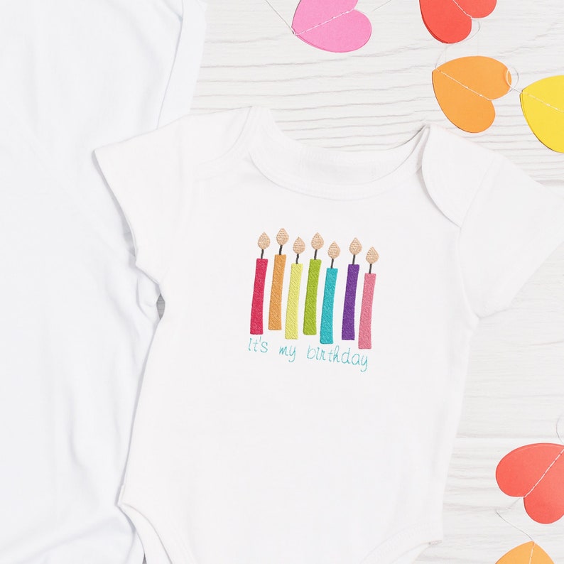 May include: White baby onesie with a rainbow of candles and the text "It's my birthday".
