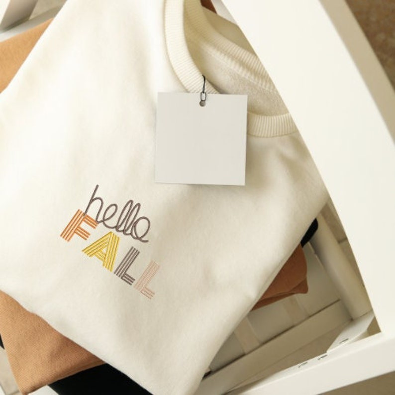 Puede incluir: Una sudadera blanca con el texto "hello fall" bordado en rayas de colores. La sudadera est&aacute; doblada y se muestra en un estante blanco.