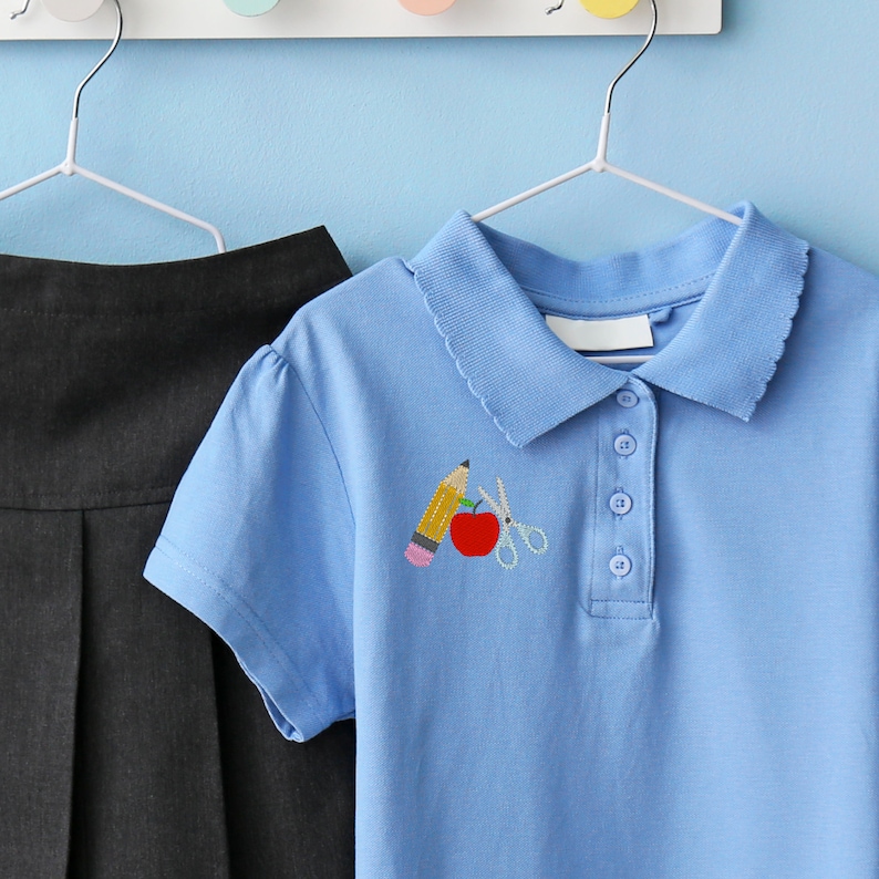 May include: A light blue polo shirt with a white collar and a small embroidered design of a pencil, apple, and scissors on the left chest.