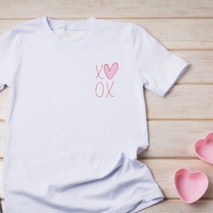 May include: White t-shirt with a red heart and the text "XOX" on the front. The shirt is lying on a wooden surface.