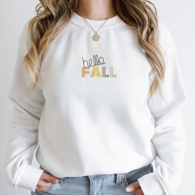 Puede incluir: Sudadera blanca con el texto "hello fall" en rayas marrones, amarillas y naranjas.
