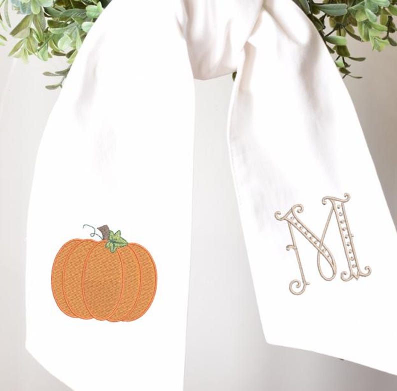 Peut inclure: Serviette de cuisine blanche avec une broderie d'une citrouille orange et la lettre "M". La citrouille a une tige et une vrille vertes. La lettre "M" est dans une police d&eacute;corative.