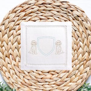 May include: White square linen cocktail napkin with a light blue shield design and two embroidered dog illustrations. The napkin has a decorative stitched border and is placed on a woven wicker placemat.