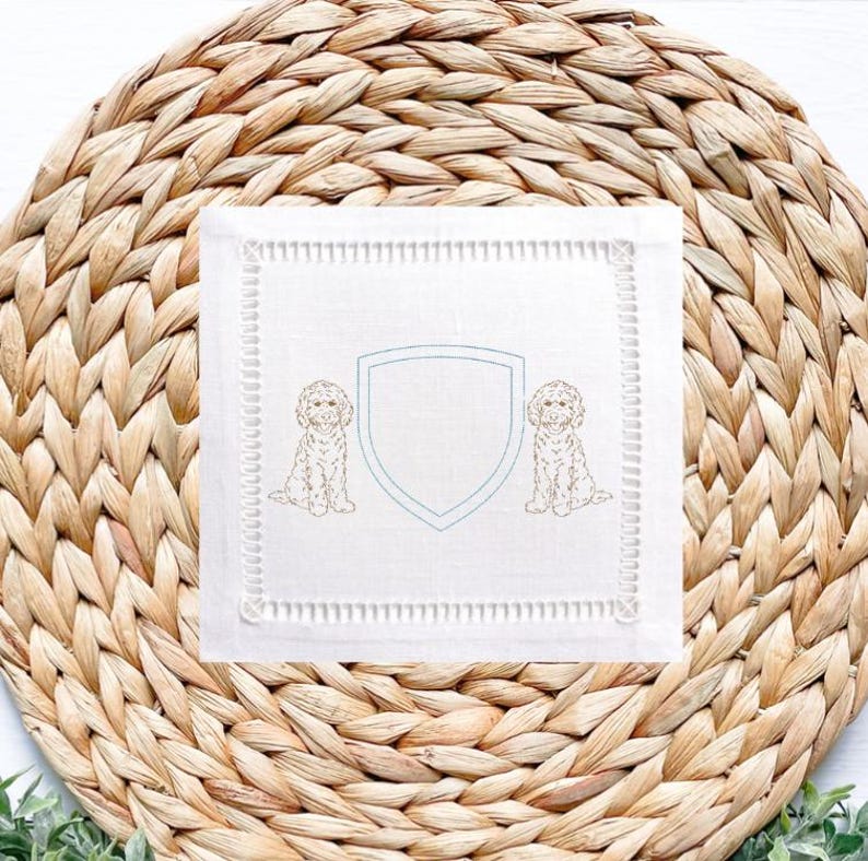 May include: White square linen cocktail napkin with a light blue shield design and two embroidered dog illustrations. The napkin has a decorative stitched border and is placed on a woven wicker placemat.