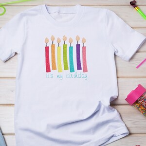 May include: White t-shirt with a rainbow of candles and the text "It's my birthday" embroidered on the front.