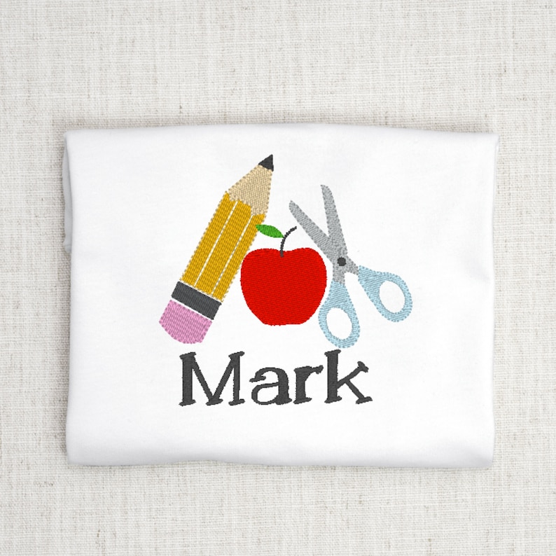 May include: A white t-shirt with an embroidered design of a red apple, a yellow pencil, and blue scissors. The name "Mark" is embroidered below the design.