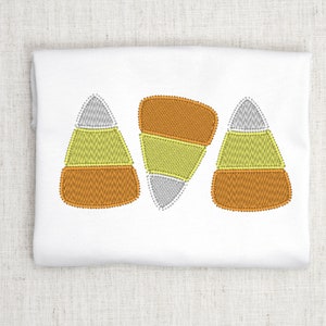May include: Three embroidered candy corn designs in orange, yellow and white on a white fabric background.