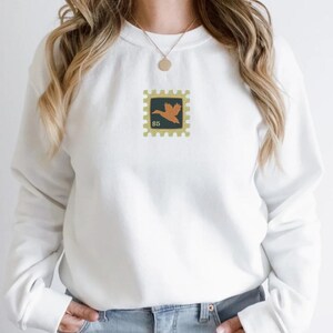 May include: White sweatshirt with a small embroidered design of a brown bird on a yellow stamp with the text "$5" on it.