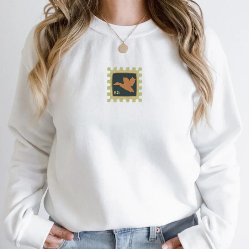 May include: White sweatshirt with a small embroidered design of a brown bird on a yellow stamp with the text "$5" on it.