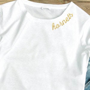 Op de afbeelding: Wit T-shirt met het woord "hornets" geborduurd in goud op de linker schouder.