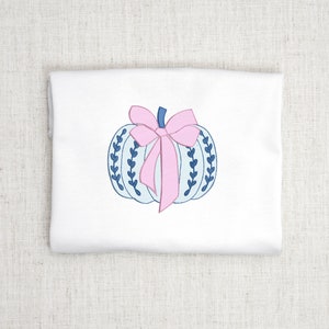 May include: A white t-shirt with a blue and white pumpkin with a pink bow. The pumpkin has a blue stem and blue leaves.