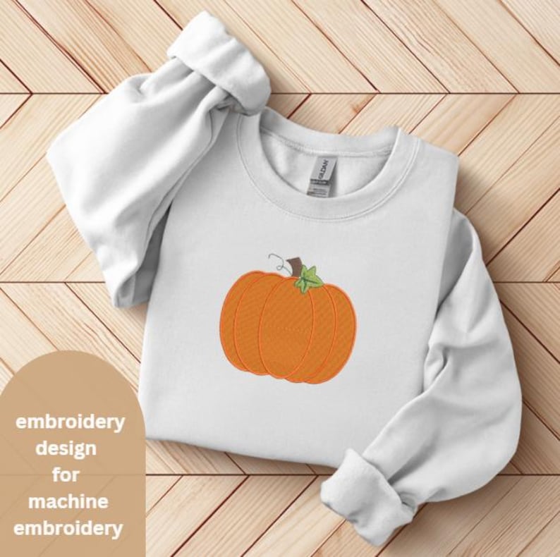 Peut inclure: Sweat-shirt blanc avec une broderie de citrouille orange et de tige verte. Le motif est centr&eacute;. Le texte "embroidery design for machine embroidery" est dans le coin inf&eacute;rieur gauche.