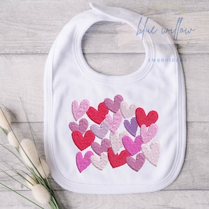 Puede incluir: Babero blanco para bebé con un diseño de corazones bordados en rosa, rojo y morado. El babero tiene un logotipo de bordado de sauce azul con el texto "Blue Willow Embroidery".