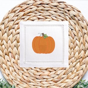 May include: A white square linen coaster with an embroidered orange pumpkin design. The coaster has a decorative stitched border and is set on a woven placemat. The pumpkin has a green stem and is a fall-themed item.