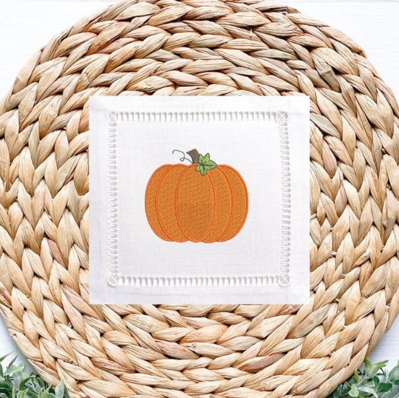 Peut inclure: Un dessous de verre carr&eacute; en lin blanc avec une broderie de citrouille orange. Le dessous de verre a une bordure cousue d&eacute;corative et est pos&eacute; sur un dessous de plat tiss&eacute;. La citrouille a une tige verte, un article sur le th&egrave;me de l'automne.