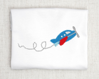 Op de afbeelding: Een wit gevouwen kledingstuk met een geborduurd ontwerp van een blauw vliegtuig met een rode vleugel, een grijze propeller en een lichtblauwe staart. Een grijs gestikt spoor volgt het vliegtuig in een lussenpatroon over de stof.