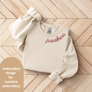 Könnte beinhalten: Ein cremefarbener Pullover mit einem roten Stickmuster, auf dem vorne "Panthers" steht. Der Pullover liegt flach auf einer Holzoberfläche.