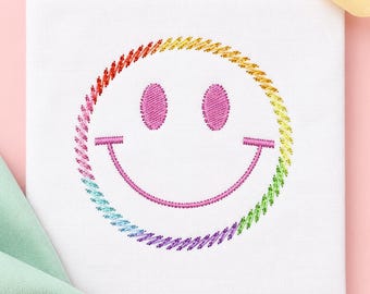 May include: A square white fabric panel featuring an embroidered smiley face design. The circular outline is stitched in a gradient of rainbow colors, while the two oval eyes and the curved mouth are embroidered in a solid bright pink thread.
