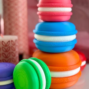 Puede incluir: Una pila de macarons coloridos con relleno blanco. Los macarons son rosa, azul, naranja y verde.
