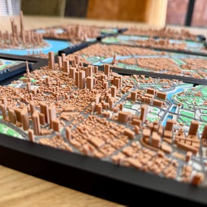 May include: A 3D printed model of a city skyline, with buildings and streets rendered in detail. The model is made of brown plastic and is mounted on a black frame.