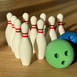 May include: A set of white bowling pins with red stripes, arranged in a triangular formation. Two bowling balls, one green and one blue, are in the foreground.