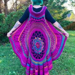 May include: A vibrant, handmade crochet vest in shades of fuchsia, purple, and rainbow colors. The vest features a large, openwork mandala design on the back and a hooded neckline. The garment is worn outdoors on a grassy lawn.