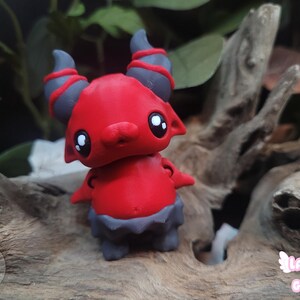 May include: A red figurine of a cartoon demon with grey horns and a grey skirt-like base. The figurine has large, white-pupiled eyes and small arms. The background features wood and foliage, with the text "Layers in Green".
