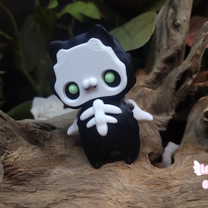 May include: A small black and white cat figurine with a skeleton design. The cat has green eyes and is wearing a black cloak. The figurine is sitting on a piece of driftwood.