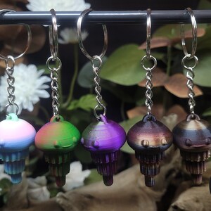 May include: Five vibrant jellyfish keyrings suspended from a metal bar. Each keyring showcases a distinct colour gradient, such as blue, green, purple, and brown. The keyrings have a detailed, layered design and are attached to silver chains and key rings.