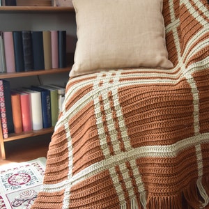 Op de afbeelding: Een gehaakt deken met een bruin en wit geruit patroon. Het deken heeft franjes aan de randen en is over een stoel gelegd met een beige kussen. De tekst "traveling hook designs" is zichtbaar in de rechterbenedenhoek van de afbeelding.