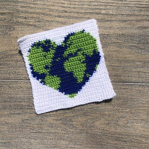 May include: A white crocheted square with a green and blue heart-shaped design depicting the Earth.