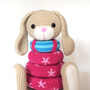 May include: A crocheted beige bunny toy with long ears and a pink nose. The bunny is wearing a stack of pink and blue crocheted rings with pink flower accents.