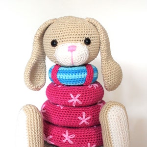May include: A crocheted bunny toy with a pink nose and brown eyes. The bunny is wearing a stack of pink and blue crocheted rings.