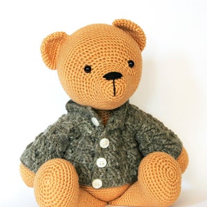 May include: A crocheted teddy bear wearing a green cardigan with white buttons. The bear is sitting with its legs crossed and has a brown nose and black eyes.