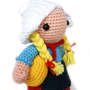 May include: A crocheted doll wearing a white cap, a blue and pink shirt, a red scarf, a dark blue skirt, and a yellow bag. The doll has yellow braided hair with red and yellow tassels.