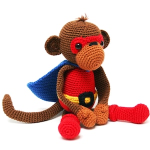 May include: A brown crocheted monkey wearing a red and yellow superhero costume with a blue cape. The monkey has red eyes and a brown nose.