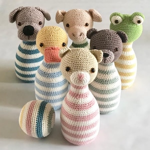 Könnte beinhalten: Eine Sammlung handgefertigter Bowlingkegel und ein Ball aus Häkelarbeit. Die Kegel sind in Form von Tieren gestaltet, darunter ein Hund, ein Schwein, eine Ente, eine Katze, ein Bär und ein Frosch. Jeder Kegel hat einen gestreiften Körper in verschiedenen Farben. Der Ball ist ebenfalls gestreift.
