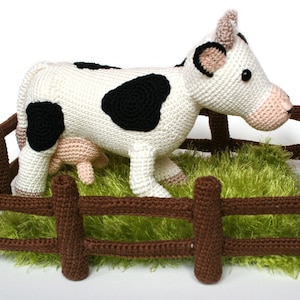 May include: A crocheted white cow with black spots standing in a brown wooden fence with green grass inside.