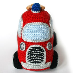 May include: A red crocheted fire truck toy with a blue light on top, black wheels, and a gray and black grill. The toy has a light blue windshield.
