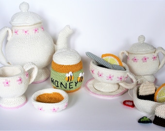 May include: A crocheted tea set with a teapot, creamer, sugar bowl, teacup and saucer, honey pot, and tea bag holder. The set is white with pink flowers and the honey pot has bees and the word "honey" on it.