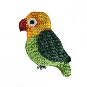 Può includere: Un giocattolo di peluche a forma di parrocchetto uncinetto con un corpo verde, una testa gialla e un becco rosso. Il giocattolo ha un occhio bianco ed è fatto con un filato morbido e testurizzato.